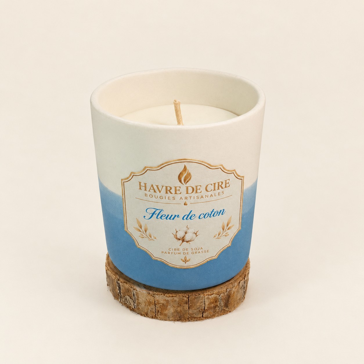 Bougie Havre de Cire Fleur de coton en cire de soja, pot en céramique blanc et bleu posé sur un rondin de bois, sur fond crème